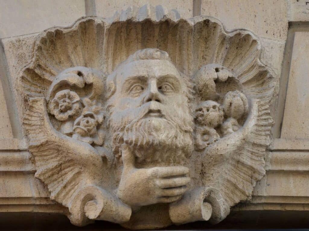 Stone carving of a bearded man's face surrounded by floral designs on a building facade in Le Marais, Paris.