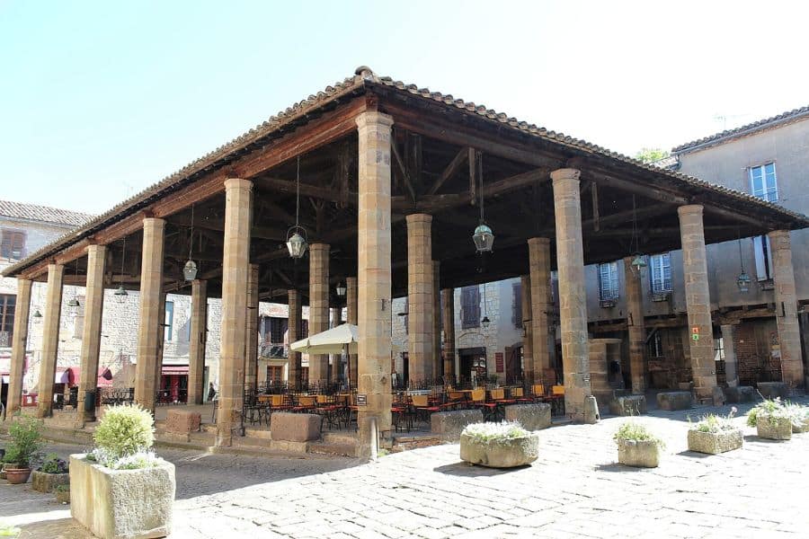 Open-air market in Cordes-sur-Ciel, featuring a large wooden-roofed structure with stone columns and hanging lanterns, surrounded by old stone buildings and cobblestone streets. A charming setting that captures the historical ambiance of France's favorite villages.