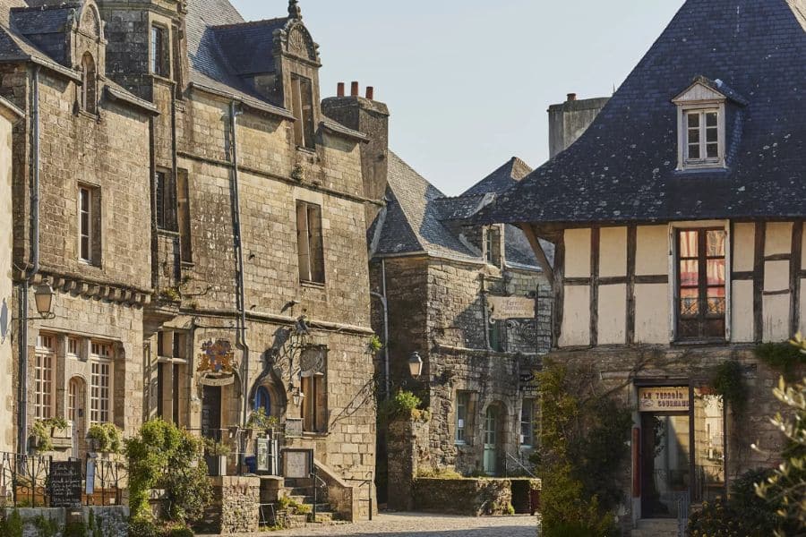 Stone buildings and half-timbered façades line a narrow street in Rochefort-en-Terre, with vintage signage and slate roofs under soft sunlight. This Breton gem is beloved as one of France's favorite villages.
