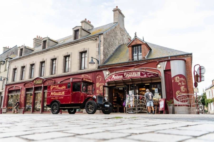 Vintage red delivery truck parked in front of Maison Gosselin, a gourmet grocery in Saint-Vaast-la-Hougue with deep burgundy storefronts and classic typography. This Normandy coastal town offers historic character among France’s favorite villages.