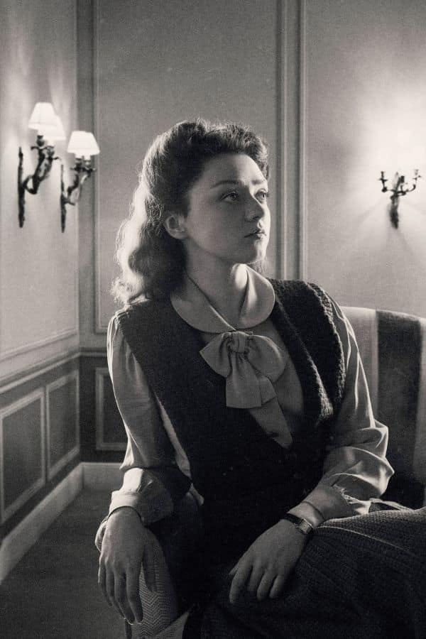 Catherine Dior gazes thoughtfully to the side in a softly lit room, wearing a blouse with a bow and a vest. The elegant setting evokes the postwar era in France.
