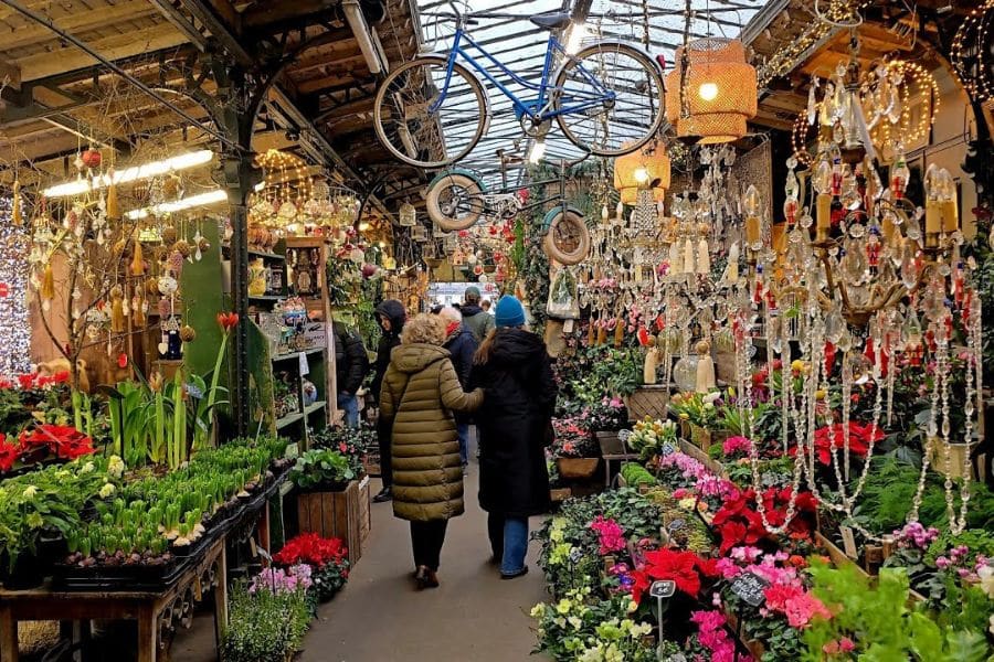 Covered flower market aisle filled with pink and red blooms, hanging chandeliers, and two shoppers walking beneath vintage bicycles suspended from the ceiling. This busy indoor scene captures the layered color and charm often associated with markets in Paris.
