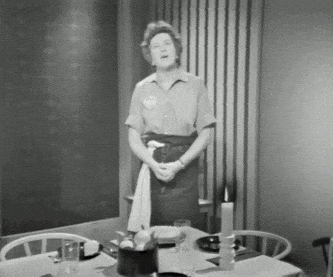Black and white still of a woman standing beside a formally set dining table with candles, plates, and folded linens, as if explaining proper table manners. It supports a discussion of French etiquette by showing a traditional, orderly dining setting and a poised host.