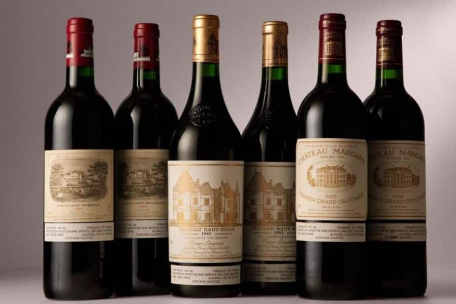 Six bottles of red wine stand in a row against a plain background with cream and gold labels facing forward. This image is useful for Burgundy vs Bordeaux content because it suggests comparing prestigious bottles, label styles, and producers from each region.