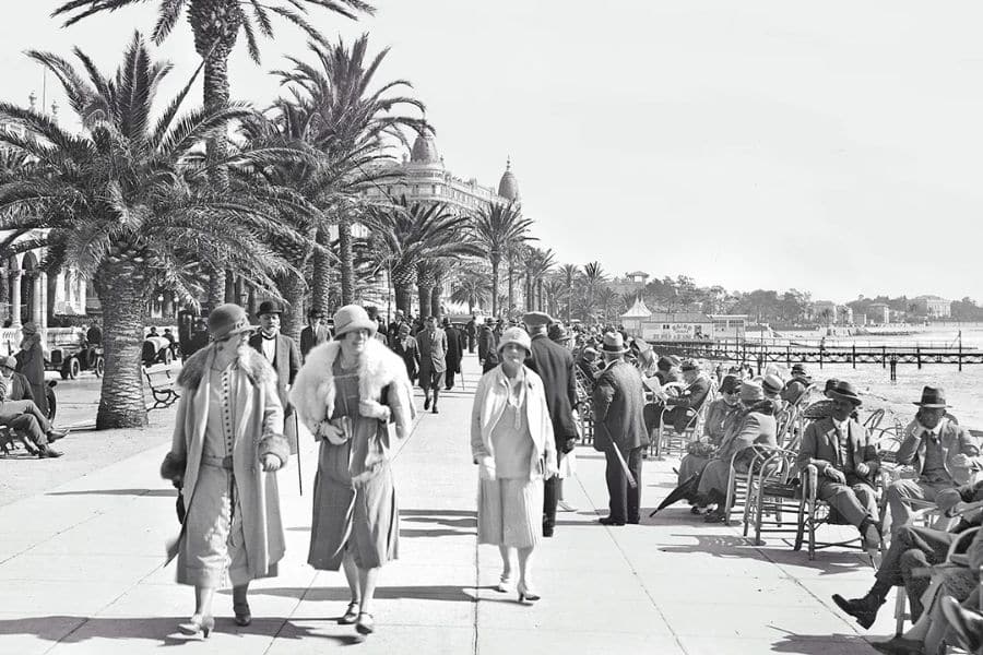 Black and white photo of people strolling along a palm tree lined seaside promenade on the French Riviera during the 1920s. Women in cloche hats and fur trimmed coats walk beside rows of seated visitors overlooking the waterfront.