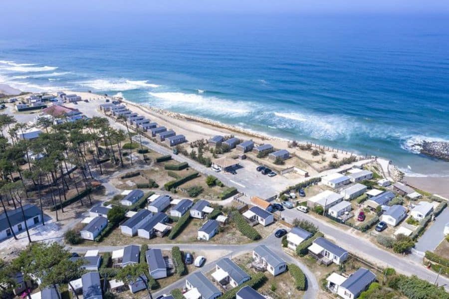 Aerial view of a beachfront camping resort in France with rows of mobile homes and cabins beside the ocean. Waves roll onto the sandy beach next to the coastal campsite on a sunny day.