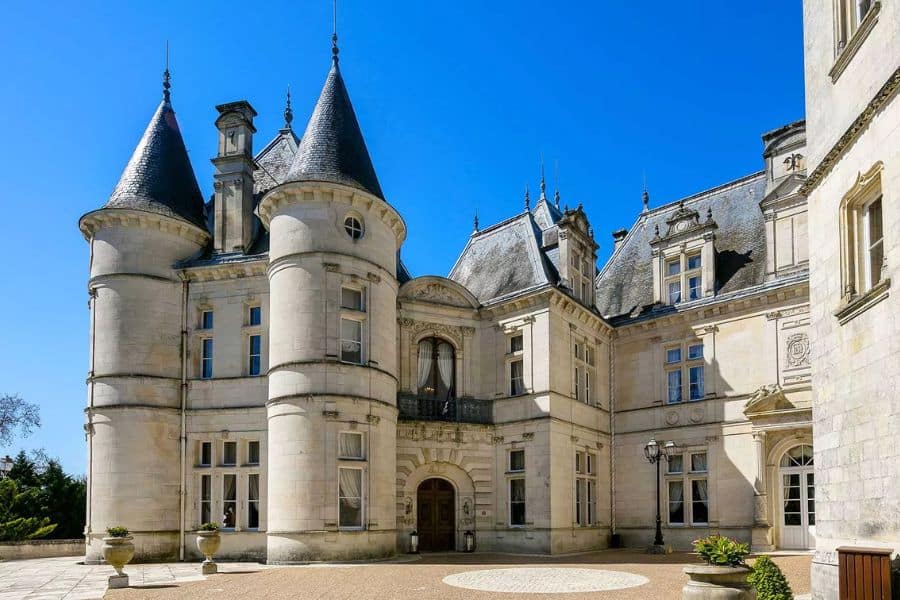 Élégant château français avec des tours rondes en pierre, des toits en ardoise abrupts et une architecture Renaissance ornée entourant une cour ensoleillée sous un ciel bleu vif. La grande façade met en valeur le luxe historique et le design raffiné associés à un château français classique.