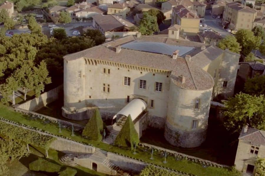 Vue aérienne d'un château historique en pierre avec des tours rondes fortifiées et des toits de tuiles entouré d'un petit village et d'un terrain paysager. Le cadre rural met l'accent sur les origines médiévales et l'architecture préservée du château français.