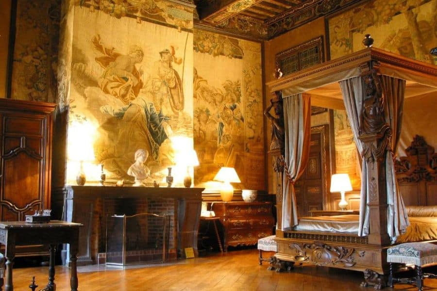 Chambre de château somptueuse dotée d'un lit à baldaquin en bois sculpté, de tapisseries murales décoratives, de meubles anciens et d'un éclairage d'ambiance chaleureux à côté d'une cheminée. L'intérieur richement détaillé reflète l'opulence et le caractère historique d'un château français de luxe.