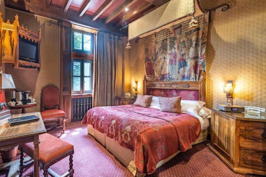Chambre d'amis confortable à l'intérieur d'un château français avec une décoration murale de style tapisserie, des meubles en bois sculpté et un couvre-lit à motifs rouges sous les poutres apparentes du plafond. Le mobilier élégant allie charme historique et confort d'un hôtel de charme.