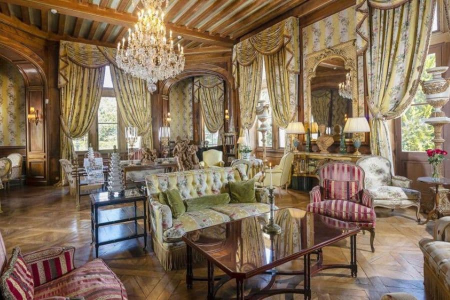 Salon luxueux à l'intérieur d'un château français comprenant des lustres en cristal, des draperies à motifs, des canapés anciens, des planchers en bois poli et des miroirs ornés. La chambre richement meublée reflète l'élégance aristocratique et le design intérieur européen classique.