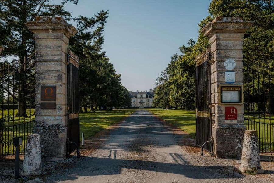 Longue allée bordée d'arbres menant à travers des portes ouvertes en fer forgé vers un château français lointain entouré de pelouses soignées et d'un parc. L'entrée formelle crée une expérience d'arrivée grandiose typique des propriétés historiques.