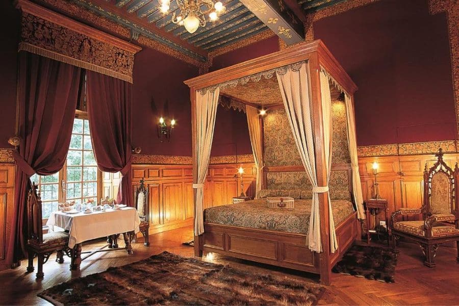Chambre château historique avec un grand lit à baldaquin, des murs rouge foncé, des boiseries sculptées, de lourds rideaux et des sièges anciens éclairés par un éclairage chaleureux de style bougie. L'intérieur richement décoré évoque l'atmosphère royale d'un ancien château français.