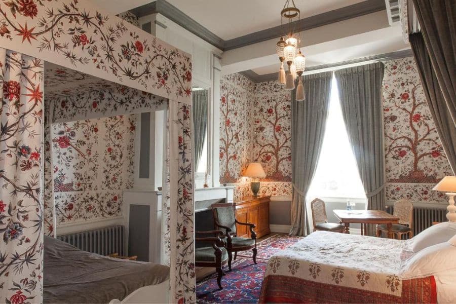 Chambre d'amis confortable à l'intérieur d'un château français avec une décoration murale de style tapisserie, des meubles en bois sculpté et un couvre-lit à motifs rouges sous les poutres apparentes du plafond. Le mobilier élégant allie charme historique et confort d'un hôtel de charme.