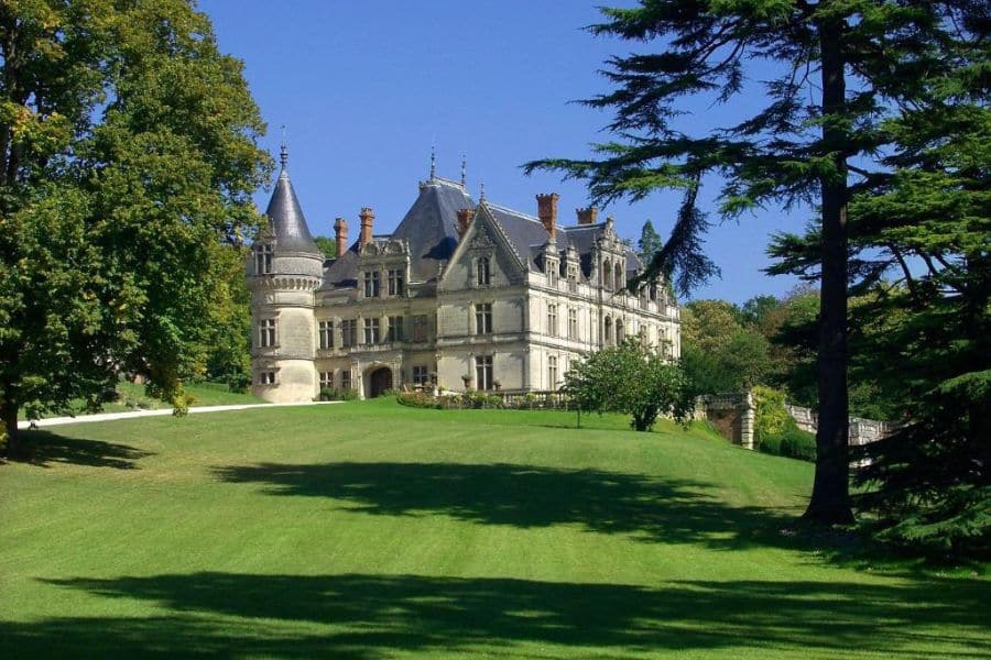 Grand château français entouré de vastes pelouses vertes, d'arbres matures et d'un parc paysager sous un ciel bleu clair. Le majestueux manoir présente des tours pointues, des pierres ornées et une architecture classique de style vallée de la Loire.