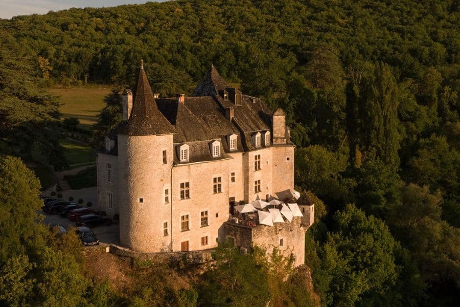 Château en pierre avec de hautes tours à tourelles perchées au-dessus d'une colline boisée au coucher du soleil, avec terrasse extérieure donnant sur la campagne environnante. L'emplacement isolé et l'architecture historique créent un cadre spectaculaire ressemblant à un château.